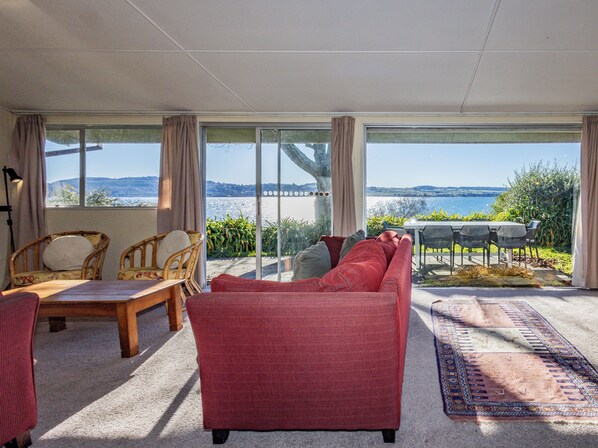 Un salon spacieux doté d’une grande fenêtre offrant une vue sur la mer et les collines. On y trouve un canapé rouge, une table basse en bois et un coin repas avec des sièges extérieurs.