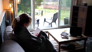 A person sitting on a couch reading a book in a room with a large window, a wooden table, and a laptop. - Smith Lake Farm (Black Creek)
