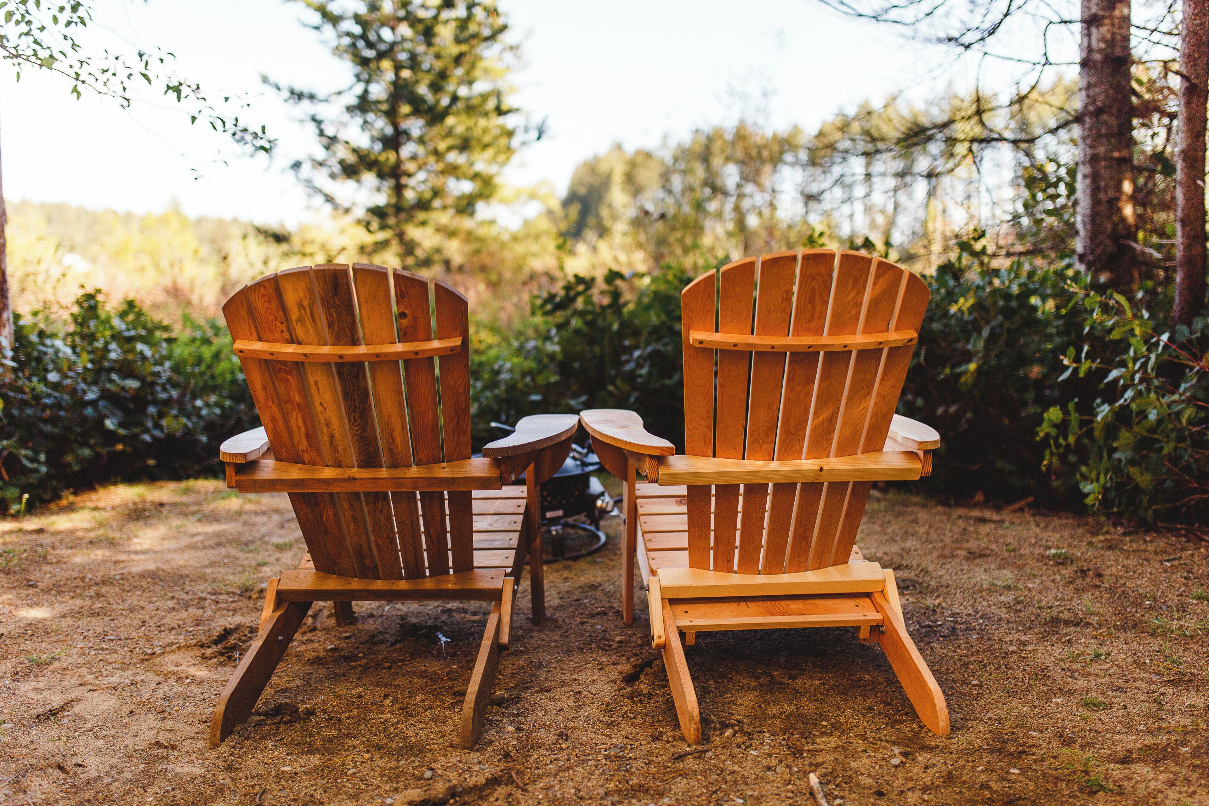 Two wooden Adirondack chairs with a small round table between them, set on a dirt ground with greenery in the background.