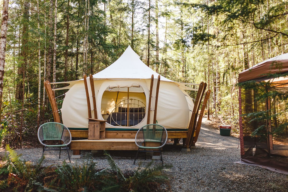 A tent with a circular entrance, surrounded by a forest. There are two chairs in front of the tent.
