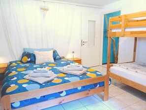 Un dormitorio con una cama litera, una cama con cubrecama estampado, una mesita de noche con lámpara y una ventana con cortinas.