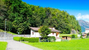 A rural setting with a paved path leading to a white building with a red roof, surrounded by lush greenery and a mountain in the background.