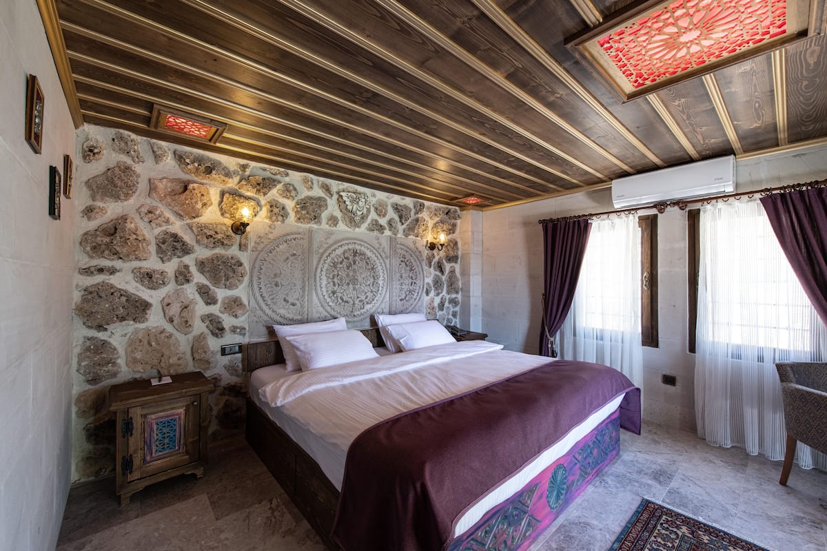 A bedroom with a stone wall, wooden ceiling, a bed with white linens, a nightstand, and a chair.