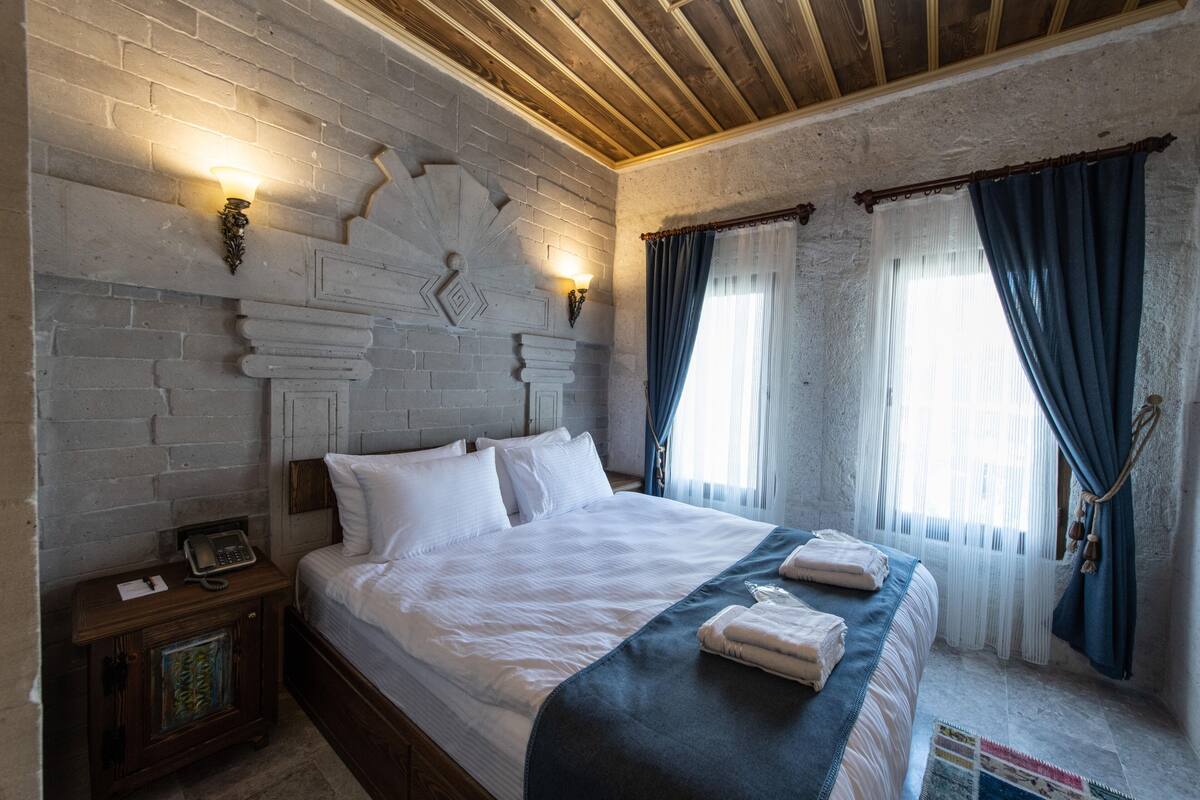 A bedroom with a brick wall, a wooden ceiling, a bed with white linens, a nightstand with a phone, and blue curtains.