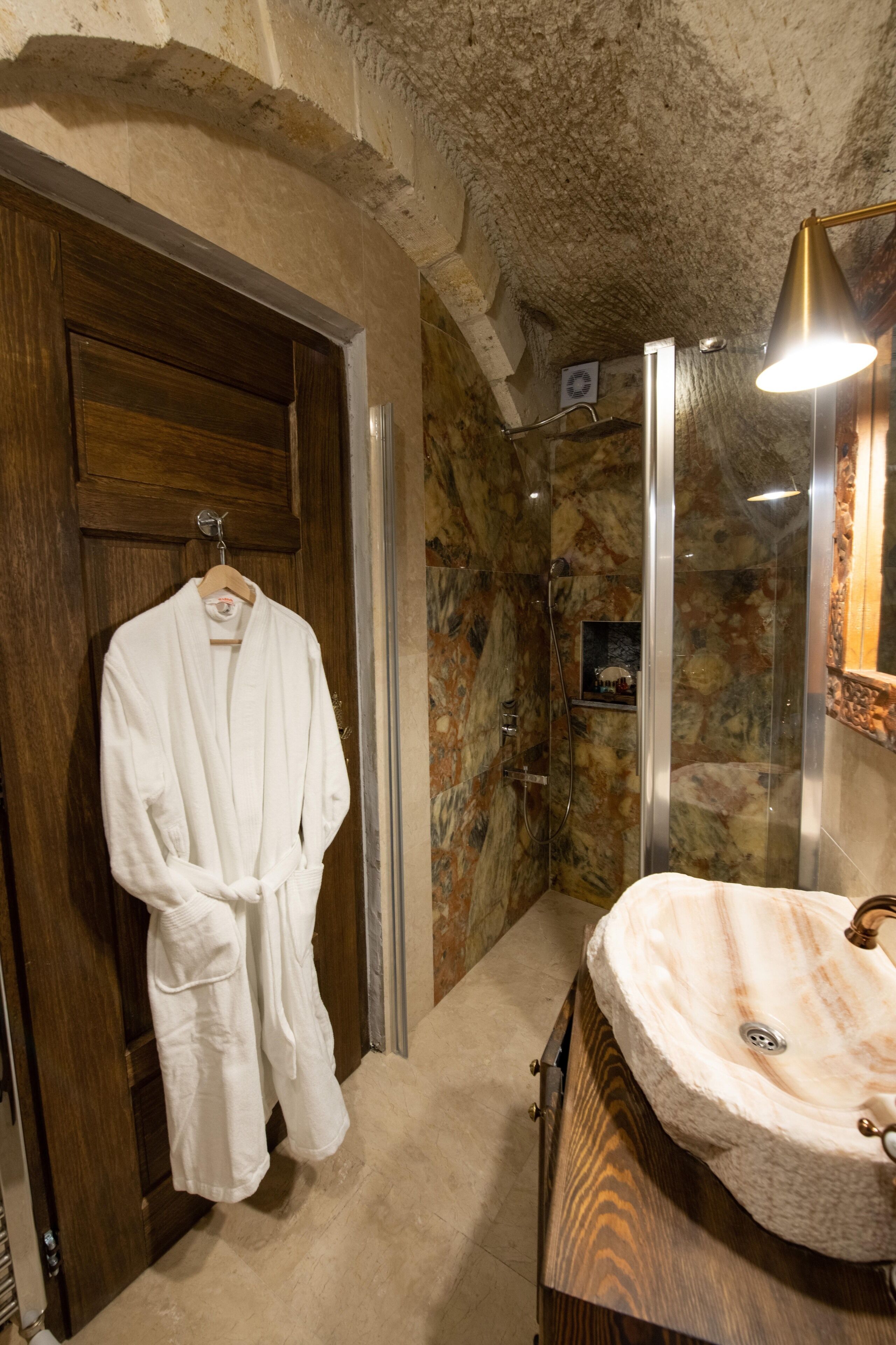 A bathroom with a marble wall, a wooden door, a white robe hanging, a glass shower enclosure, and a sink with a marble countertop.