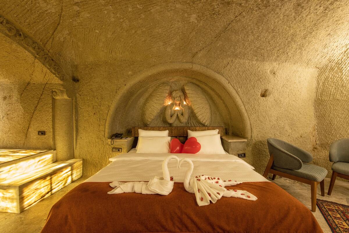 A room with a stone ceiling, a bed with a brown bedspread, two swan-shaped towels, and a pair of red heart-shaped pillows.