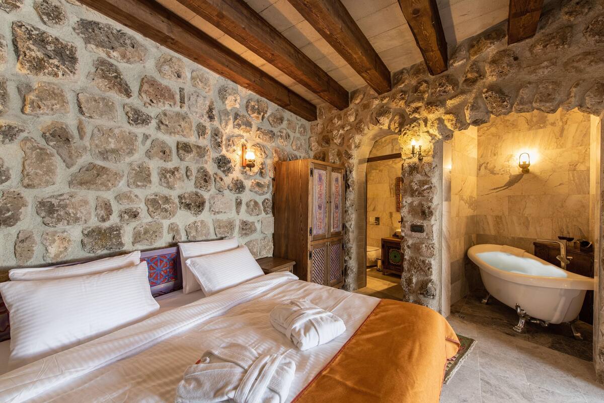 A stone-walled bedroom with a large bed, a wooden ceiling, and a vintage-style bathtub.