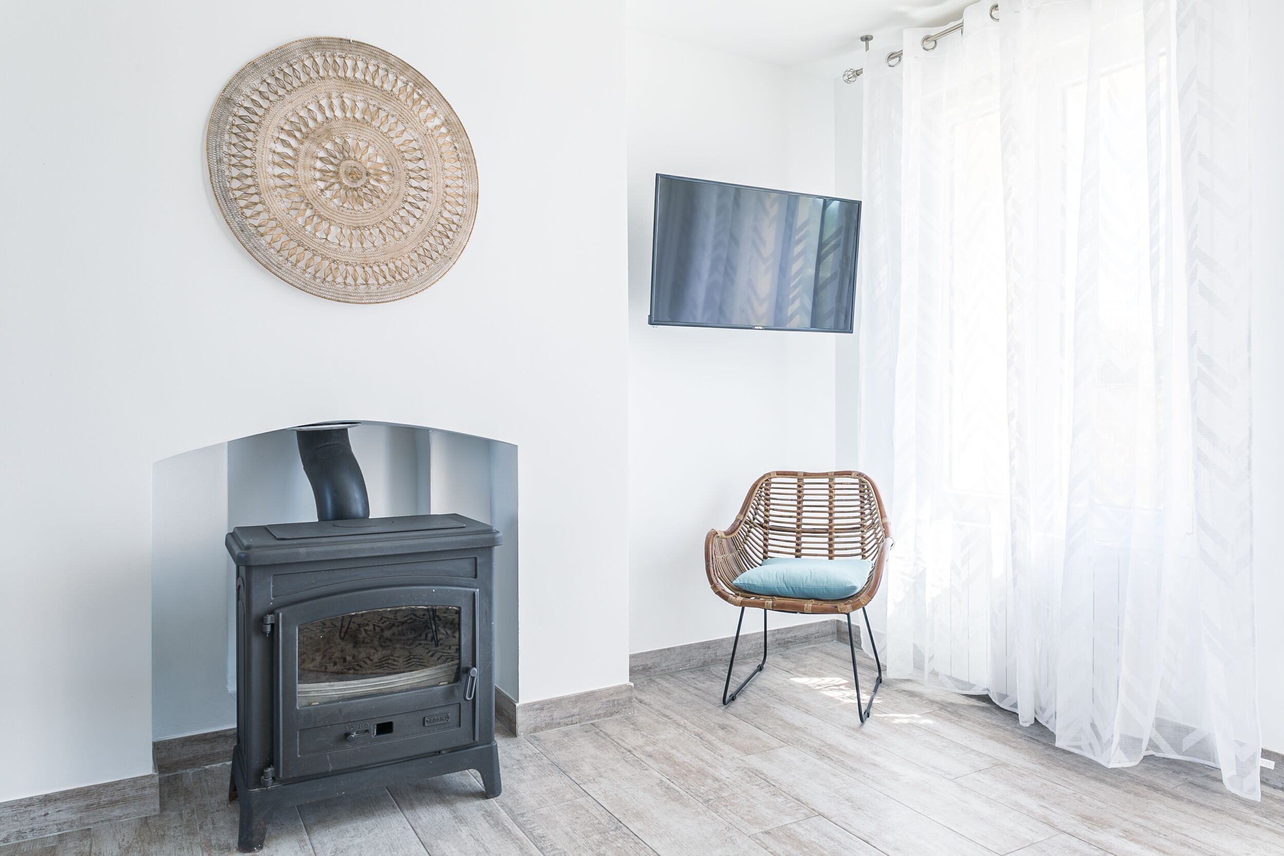 Een kamer met een houtkachel, een geweven wanddecoratie, een flatscreen televisie, een rieten fauteuil en lichte gordijnen.