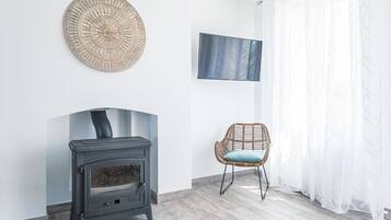 Een kamer met een houtkachel, een geweven wanddecoratie, een flatscreen televisie, een rieten fauteuil en lichte gordijnen.