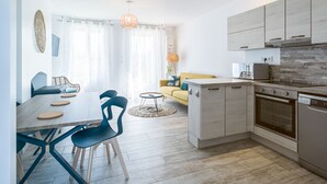 A modern kitchen with wooden cabinets, a built-in oven, and a dishwasher. There is a dining area with a table and chairs, and a yellow sofa in the background.