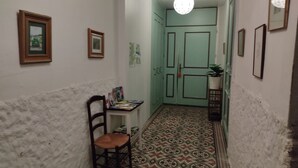 Un pasillo estrecho con piso estampado, una silla, una mesita con libros y una puerta verde.