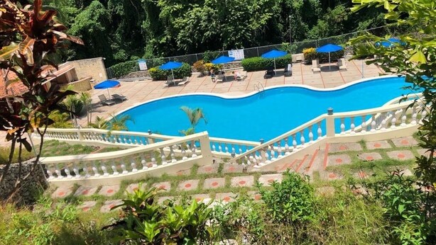 Una piscina con barandilla blanca, rodeada de follaje y algunas sillas de descanso.