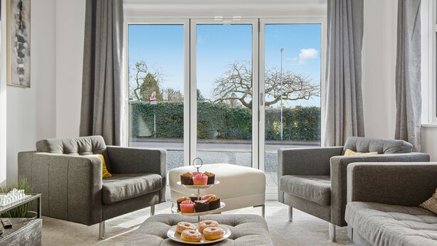 A modern living room with grey sofas, a coffee table, and a view of a street and greenery through large windows. - Luxury 2 Bedroom Apartment in Kettering (Kettering)