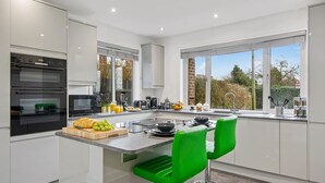A modern kitchen with a central island, two green bar stools, and a view of the outdoors through large windows. - Luxury 2 Bedroom Apartment in Kettering (Kettering)
