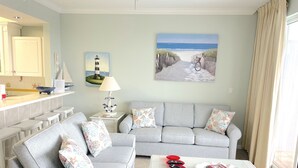 Un salon avec un canapé gris, une table basse en verre et un tableau représentant une scène de plage.