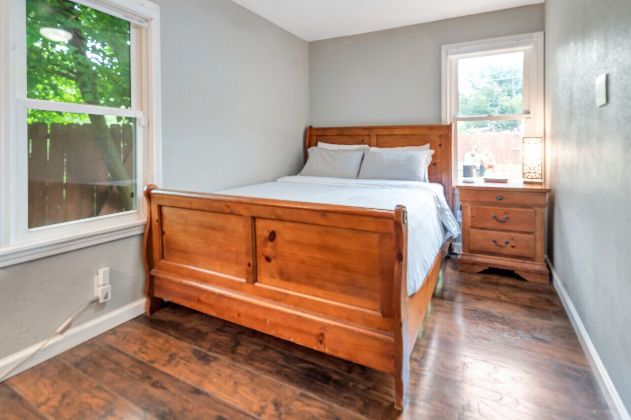 A bedroom with a wooden bed, a nightstand, and a window with a view of a backyard.