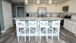 Una cocina moderna con armarios blancos, un refrigerador de acero inoxidable, un microondas y una cafetera. Hay tres taburetes blancos en la isla.