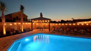 Una piscina con cubierta de madera y sillas de descanso, rodeada por una valla de madera y luces colgantes.