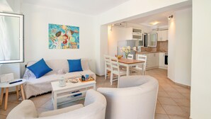 Ein modernes Wohnzimmer mit einer Couch, Sesseln und einem Esstisch. Im Hintergrund befindet sich eine Küche mit weißen Schränken und einem Kühlschrank.