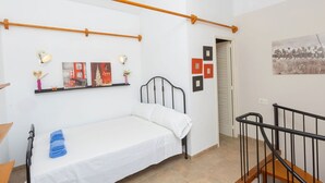 Un lit simple avec un matelas blanc et des serviettes bleues, une tête de lit en bois et un cadre en métal noir. Des appliques murales et des tableaux encadrés sont accrochés au mur au-dessus du lit.