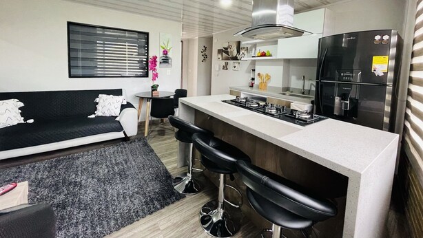 A compact kitchen with a gas stove, a refrigerator, and a dining area with a black sofa and two bar stools. - 502 Hermoso Apartamento Amoblado Movistar Arena (Bogotá)