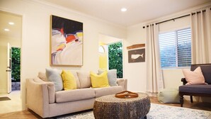 A living room with a beige sofa, yellow pillows, a woven ottoman, and a patterned rug. - Mile to Ocean! Fully Fenced Private SoCal Cottage (Oceanside)