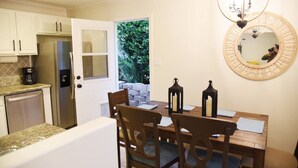 A compact kitchen with white cabinets, a stainless steel refrigerator, and a dining area with a wooden table and chairs. - Mile to Ocean! Fully Fenced Private SoCal Cottage (Oceanside)
