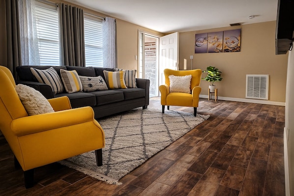 A living room with a dark sofa, yellow armchairs, a patterned rug, and a large window with blinds.