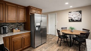 A kitchen with wooden cabinets, a stainless steel refrigerator, a coffee bar, and a dining area with a table and chairs. - Remodeled 2BR Near Gulf Beaches Fast Wi-Fi (Holiday)