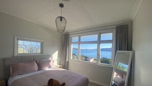 A bedroom with a bed, a nightstand, a lamp, a window with a view of the sea, and a ceiling fan. - Amazing views! 5 mins to beach (Dunedin)