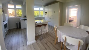 A modern kitchen with white cabinets, a central island, and bar stools. There is a dining area with a round table and chairs. - Amazing views! 5 mins to beach (Dunedin)