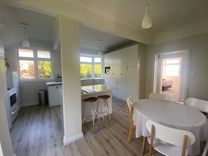 A modern kitchen with white cabinets, a central island, and bar stools. There is a dining area with a round table and chairs. - Amazing views! 5 mins to beach (Dunedin)