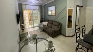 Una sala de estar con sofá, una mesa de comedor con sillas, televisor montado en la pared y un ventanal con cortinas.