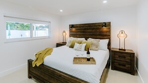 A modern bedroom with a wooden headboard, a large bed with white bedding, two bedside tables with lamps, and a window with a view of greenery. - Encanto Family, cozy patio & pool, near to everything (Miami)