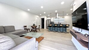 A modern living room with a sofa, a glass coffee table, and a TV. There is a kitchen area with a fridge, stove, and bar stools.