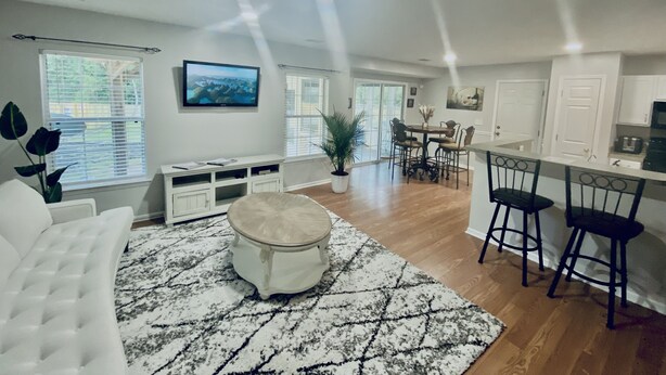 A spacious living area with a white sofa, a coffee table, and a TV mounted on the wall. There is a dining area with a table and chairs, and a kitchen with a countertop and cabinets.