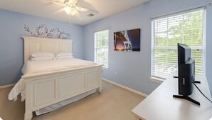 A bedroom with a white bed, a ceiling fan, a TV on a stand, and two windows with blinds.