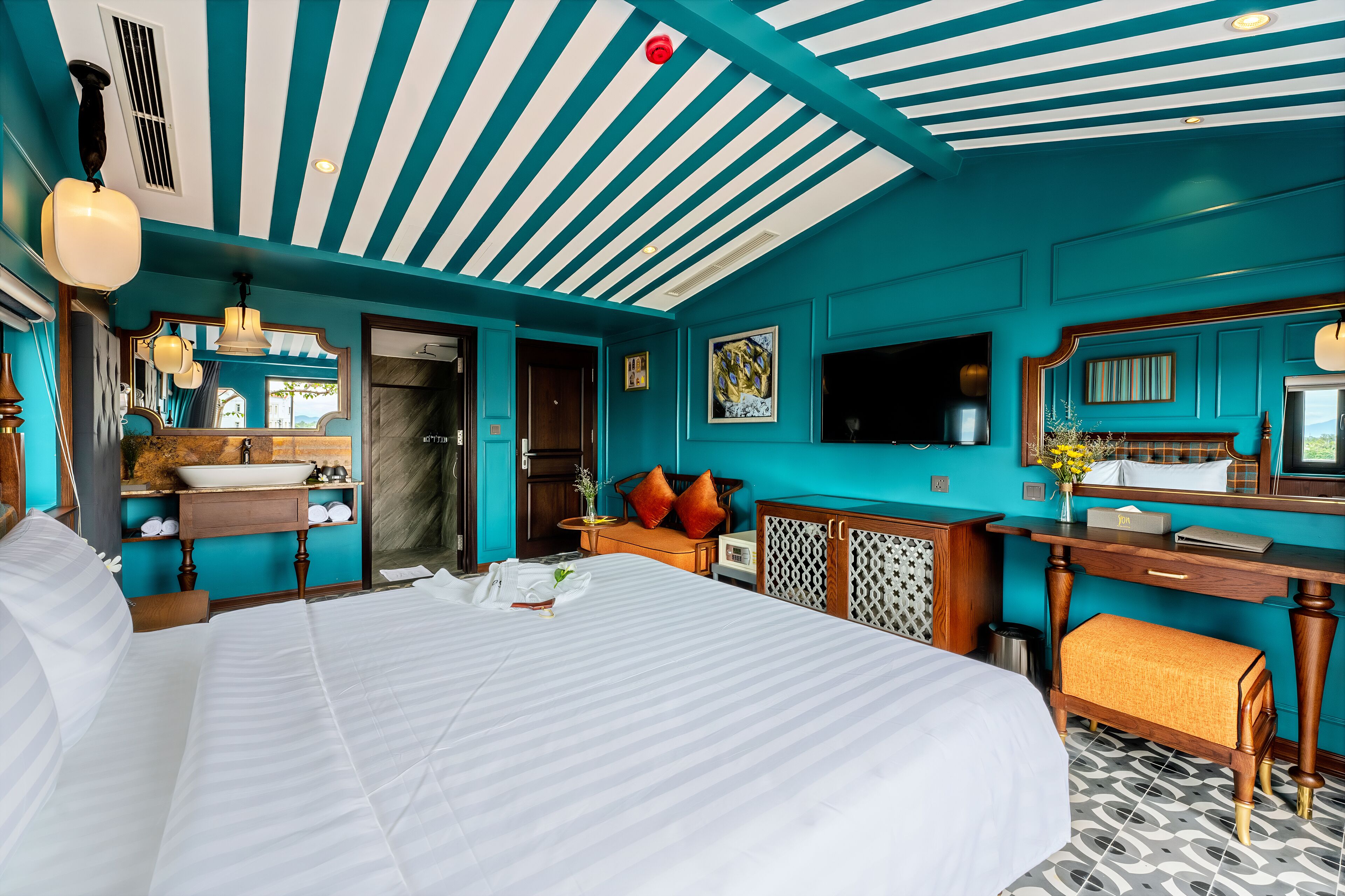 A hotel room with a large bed, a wooden desk, a bathroom with a sink and mirror, and teal walls.