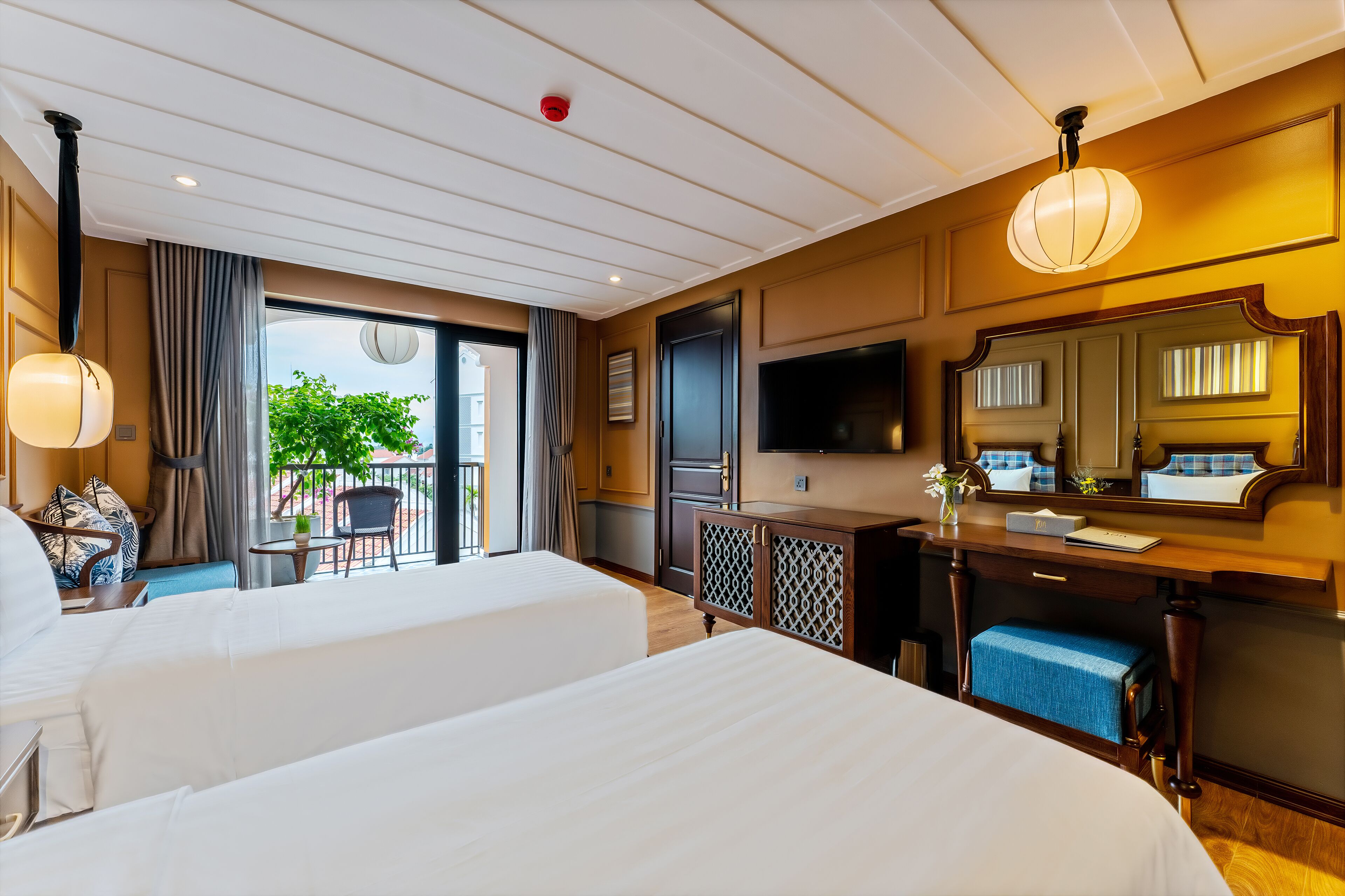 A hotel room with two beds, a TV, a desk with a mirror, and a balcony with a view of greenery.