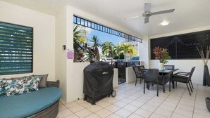 Une terrasse dotée d’un barbecue couvert, d’une table à manger avec des chaises, d’un canapé aux motifs tropicaux et d’une vue sur des palmiers et une rue.