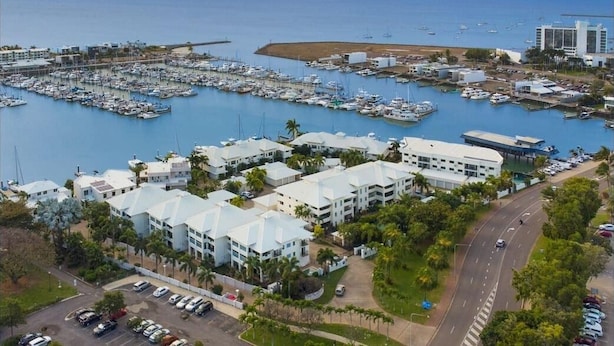Vue aérienne d’une marina avec des bateaux, un parking et des bâtiments aux toits blancs.
