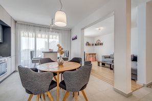Uma sala de estar moderna com uma mesa redonda de madeira, cadeiras estofadas cinzas, um sofá e uma área de jantar com uma mesa e cadeiras brancas.
