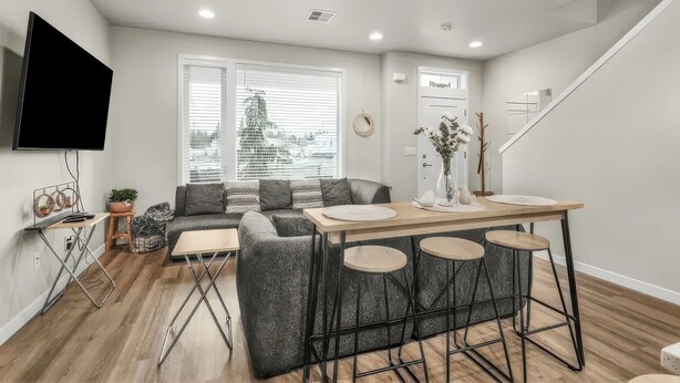 A modern living room with a sofa, a dining table with stools, a TV mounted on the wall, and a staircase. - Tranquil Townhome in Prime Location (Vancouver)