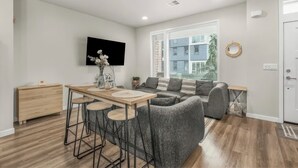 A modern living room with a sofa, a dining table with stools, a TV mounted on the wall, and a window with blinds. - Tranquil Townhome in Prime Location (Vancouver)