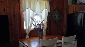 Une table de salle à manger en bois, accompagnée de deux chaises, d’une plante en pot et d’une fenêtre avec des rideaux.