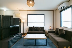 A modern living room with a flat-screen TV, a sofa, a coffee table, and a large window with blinds. - Naha Nishi EXseed (Naha)