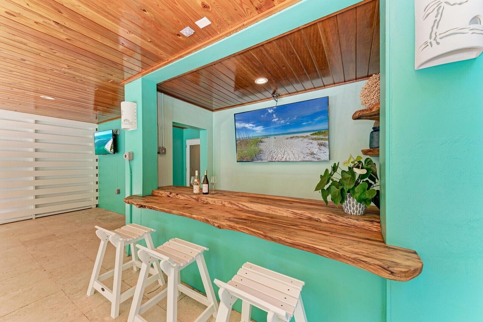 A bar area with a wooden counter, two white stools, a large framed picture of a beach, and a potted plant.