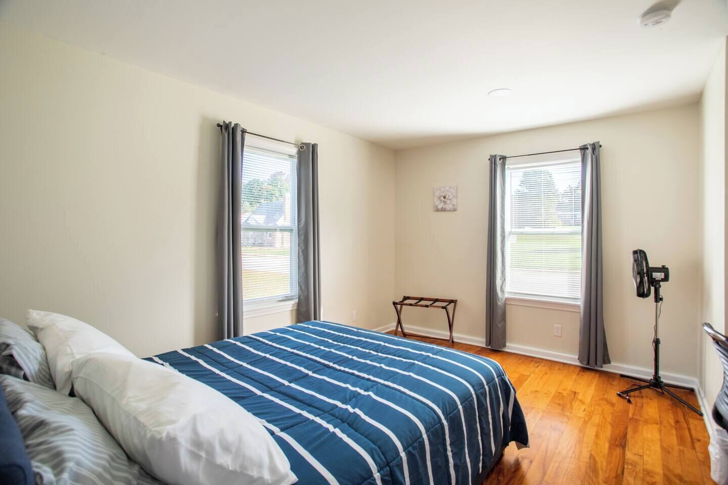 A bedroom with a bed, two windows with curtains, a standing fan, and a small table.