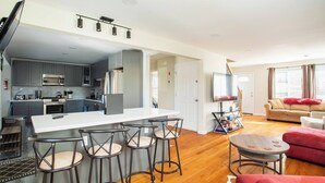 Una cocina moderna con armarios grises, una isla central con taburetes de bar, un comedor con una mesa redonda y una sala de estar con un sofá y televisor.