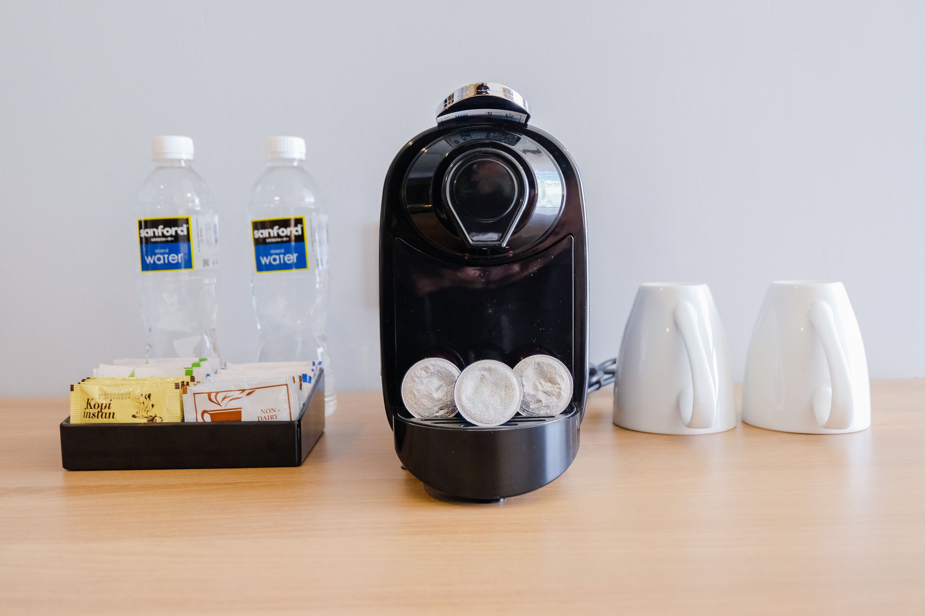 A coffee maker with three coffee cups, two bottles of water, and a tray with coffee pods and a box of instant coffee.